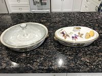 Two English china serving dishes on a kitchen counter; one round with glass lid, one oval with painted fruit and floral patterns.