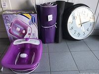 Purple foot massager with rollers in and out of box, positioned next to two yoga mats and wall clock.