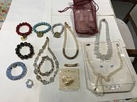 Photo showing multiple beaded bracelets in various colors including red, black, blue, turquoise, and grey, a beaded ring, a brooch in packaging, and several necklaces including a thick frosted bead necklace and a braided cord necklace.