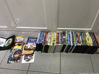 Stack of 44 children's DVD movies, cases lined up on the floor against a white door. Some discs and covers visible showing family and animated titles.