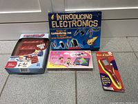 Four boxed items aligned on floor: Introducing Electronics kit, Guess Who board game, Bop It! game, and Etch A Sketch Game Pen