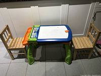 Full view of the kids activity table in plastic with blue and white drawing surface, orange side compartment, and green legs, flanked by two wooden chairs.