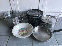 Photo showing full assortment of kitchen pots, pans and ceramic bowl arranged on floor with white cabinets in background