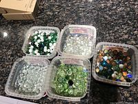 Five small plastic containers each filled with different types of marbles including clear, green, white, black, and multicolored swirled decorative marbles on a granite countertop.