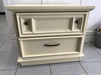 Front view of one cream-colored wooden night table showing two drawers with metal handles and decorative front panel