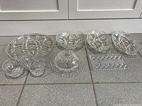 Nine pieces of clear pinwheel patterned crystal arranged on floor: large oval tray, covered butter dish, footed candy dish, two smaller oval dishes, two small round dishes, sugar bowl, and creamer