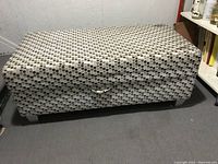 Closed view of rectangular storage bench upholstered in patterned fabric with handle on front.