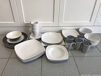 Set of white dishes arranged on a kitchen counter including plates, bowls, and mugs, some with black rims and accents.
