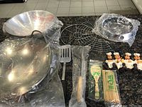 Photo showing the entire set on a dark granite countertop including a large stainless steel wok, wok stand, metal wire rack, various cooking utensils in packaging, bamboo skewers, and four chef figurines.