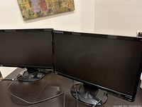 Two black widescreen BenQ monitors side by side on a dark wood surface with their display screens off and monitor stands visible. VGA cables attached.