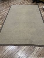 Photo of the large rectangular beige area rug with brown edging laid on wood flooring. Mild wear and small stains visible on the surface.