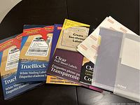 Photo of various unopened and partially used packs of TrueBlock white mailing labels, clear full sheet labels, clear business card labels, and Oxford brand items arranged on a dark surface.