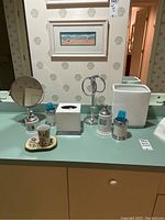 Photo showing the entire collection of white ceramic and metal bathroom accessories arranged on a countertop with mirror and wallpaper background.