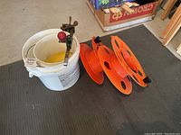 Two white paint buckets, one with brass spigot and yellow hose, and two orange wire holders with black ends.