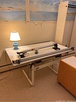 Photo of white Neolt drafting table with adjustable metal legs and attached drafting rail system next to a blue-based ceramic table lamp with white lamp shade.