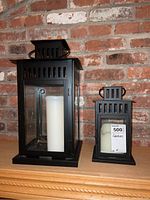 Both metal lanterns on wooden surface with brick wall background, showing white pillar candles inside and price tag on smaller lantern.