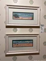 Two framed and matted prints hanging on a wall with shell-patterned wallpaper. Both prints feature beach scenes and are horizontally oriented with cream-colored frames.