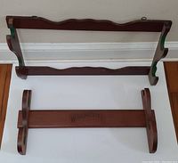 Two wooden rifle racks displayed flat on the floor, one with a carved design and green accents, the other a flat wood rack with Winchester branding.