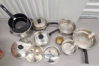 Assorted stainless steel pots and frying pans with multiple matching glass lids and one small brass basket on a gray table.