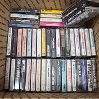 Photo of multiple cassette tapes of rock and pop artists in a cardboard box. Visible artists include Madonna, Eric Clapton, The Cars, Barry White, Rod Stewart, B.B. King, Bruce Springsteen, Gladys Knight, Boz Scaggs, George Thorogood.