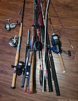 Multiple fishing poles in a pile on wooden floor, showing various handles and reels