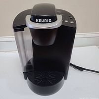 Front view of the Keurig coffee maker showing water reservoir, control buttons, power cord, and drip tray