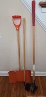 Full view of three gardening tools including a rotary edger, orange metal shovel with D-handle, and small digging shovel with wooden handles against a wall