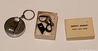 Photo showing a box with three black and one metal banjo fingerpicks alongside a round metal tape measure keychain with an attached key ring.