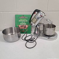 Vintage Dormeyer Silver Chef stand mixer with power cord unravelled on white table. Includes two stainless steel mixing bowls and Betty Crocker's Dinner in a Dish cookbook propped behind.