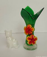 Frontal photo showing the iridescent white cat figurine sitting next to the green glass vase with orange flowers.