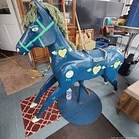 Front and side view of vintage wooden folk art horse, painted in blue with yellow heart and flower motifs, mounted on metal base, showing yarn mane and sturdy construction.
