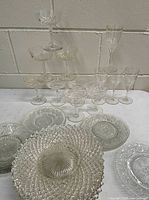 Photo showing arrangement of clear vintage glass and crystal drinkware and plates against a cinder block wall on a white table.