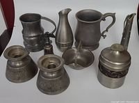 Photo showing all eight pewter items together arranged neatly on a flat surface, including candleholders, vases, lidded box, pitcher, tankard mug, and spray bottle.