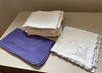 Three stacks of napkins: a stack of blue linen napkins, a stack of white embroidered cotton napkins with scalloped edges, and a larger stack of plain white damask napkins.