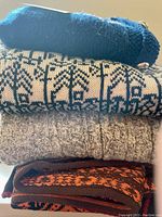 Folded stack of four sweaters showing various knit patterns and colors including navy, black and cream, light brown cable knit, and orange and black patterns.