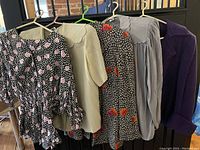 Five garments hung on hangers against railing: two floral tops, cream top, grey top and purple coat