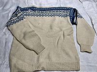 Full view of a cream wool sweater with blue geometric pattern across the shoulders and upper chest, laid flat showing long sleeves and ribbed cuffs and hem.