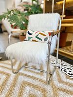 Front angle showing white faux fur covering chair, chrome frame, oak armrests, and colorful geometric pillow for scale.