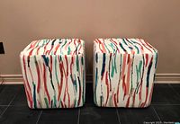 Front view of two cube-shaped ottomans covered in white fabric with colorful painted vertical stripes.