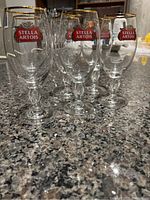 6 clear Stella Artois beer glasses with gold rim and logo arranged on granite counter.