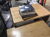 Five coffee table books stacked on a light wood table, showing front covers and spines for identification.
