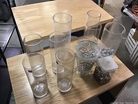 Photo of 7 glass vases on a table in a warehouse setting showing different sizes and one or two with small stones inside.