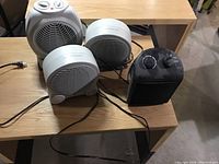 Four space heaters placed on a wooden desk in a room. Heaters vary in style and size displaying their front grills and control knobs/dials.