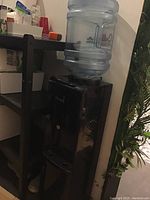 Full view of the black Primo water cooler with the 3-gallon water jug on top, set beside a black shelf and near a plant.