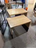 Two oak plywood retail display tables shown stacked, larger table behind smaller, displaying smooth oak finish and black lower leg accents.