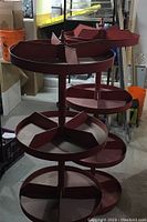 Two red round steel tiered retail display shelves with multiple divided sections, showing visible signs of wear and dust in a storage space.