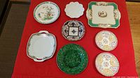 Overview of the 8 decorative antique and vintage plates laid on red cloth including pheasant, floral, Majolica leaf, scalloped Limoges and Imari-style plates