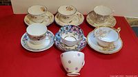 Seven assorted china teacups and six saucers arranged on a red surface. Various floral and gold detail patterns visible, including a teacup with purple polka dots.