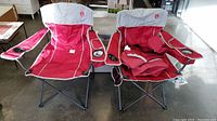 Two Coleman oversized quad camping chairs side by side, showing overall red and gray color with arm coolers and drink holders.