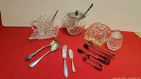 Full lot overview showing silver-plated sugar tongs, butter spreaders, demitasse spoons, etched glass sugar bowl with silver lid, antique crystal box, mint sauce and mustard bowls.